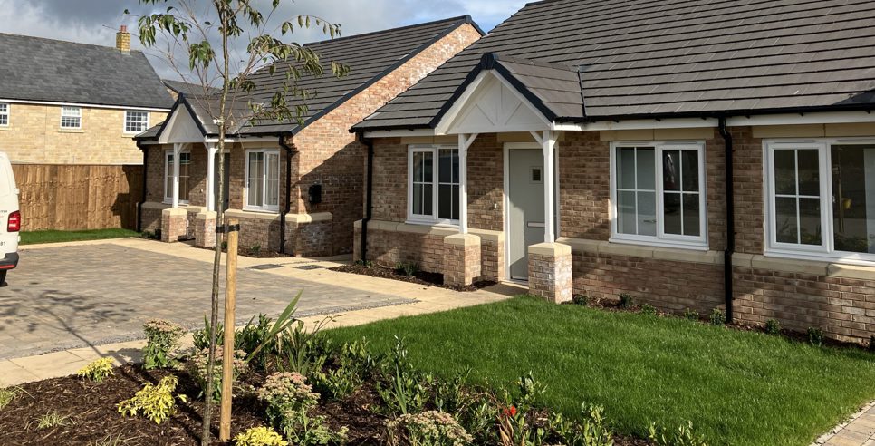 two newly built bungalows with light bricks and slate roofs. They have a small garden and driveway.