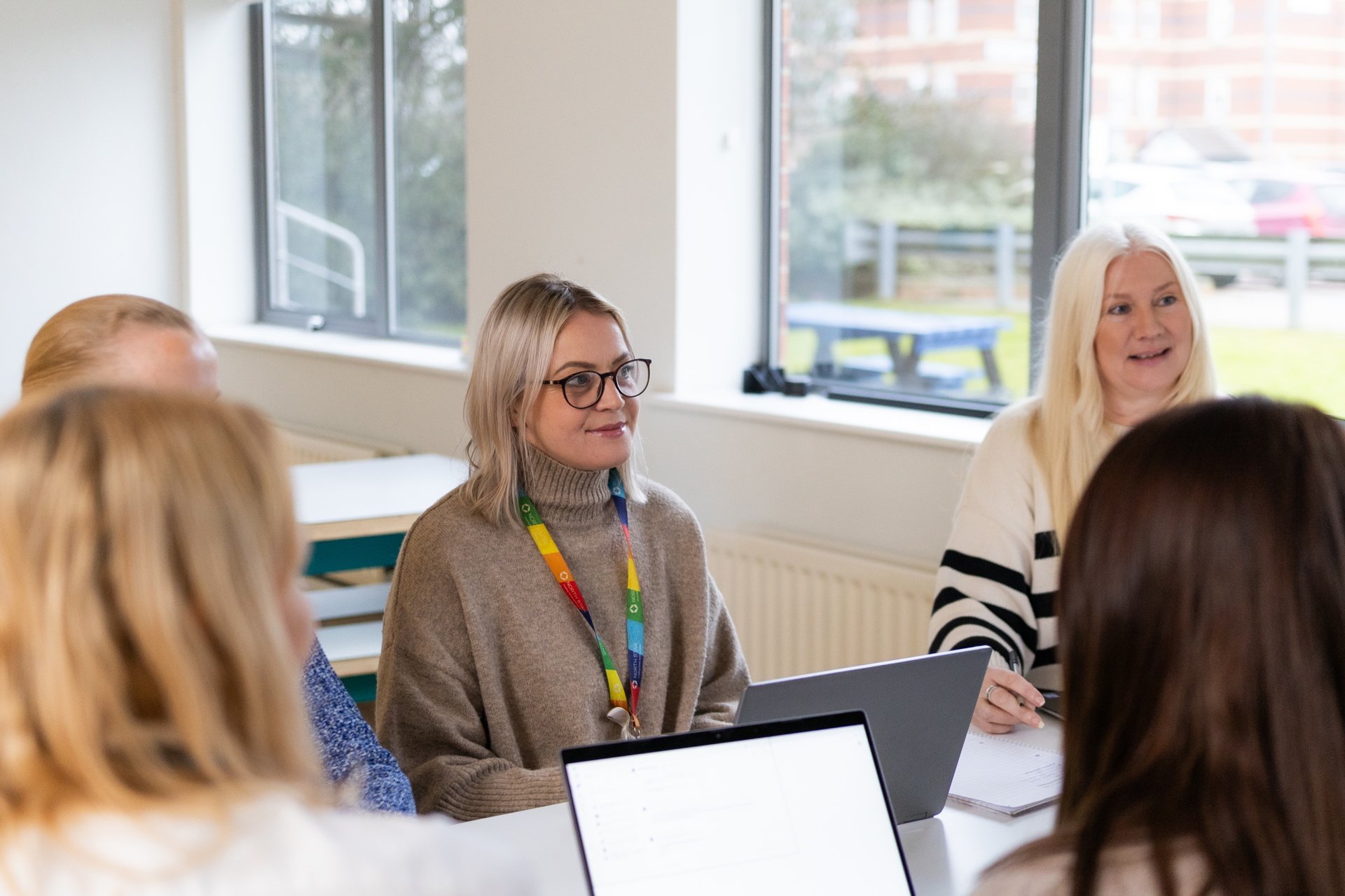 A group of women in a bright office meeting room, engaged in discussion around a table with laptops and notebooks. One woman in a beige turtleneck sweater and rainbow lanyard is listening attentively, while another woman with long blonde hair and a striped sweater is smiling. Large windows in the background let in natural light and reveal a view of parked cars and a picnic bench outside.