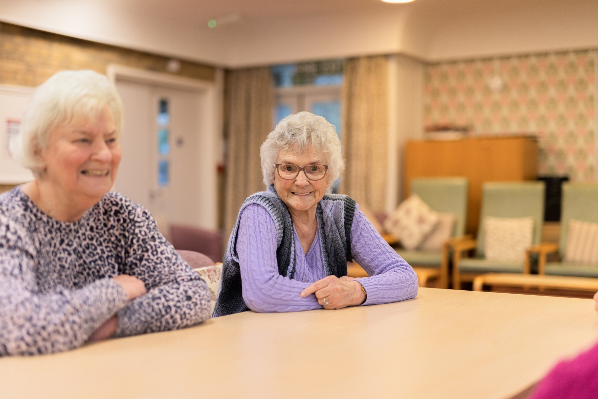 Two elderly ladies sat at a table smiling in a cosily furnished room. 