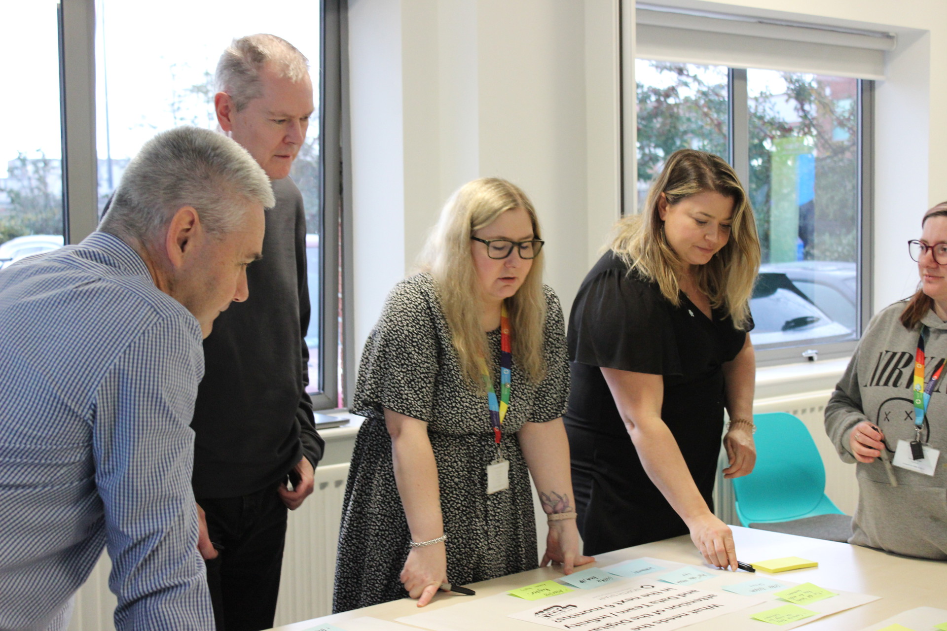 A group of five people gathered around a table in a well-lit office space, engaged in a collaborative activity. They are reading and discussing a large sheet of paper covered with text and surrounded by colorful sticky notes. Two women are pointing or writing, while the others observe attentively. Windows in the background show parked cars and trees outside.
