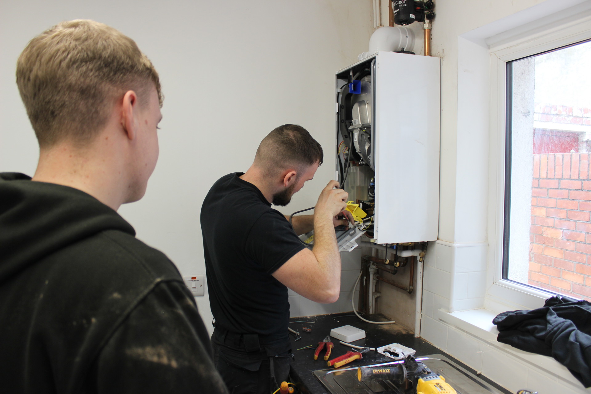 Two men in workwear are servicing a wall-mounted boiler. One man is actively working on the open boiler using tools, while the other observes closely. Various tools are scattered on the countertop below, and a window to the right lets in natural light, showing a red brick wall outside
