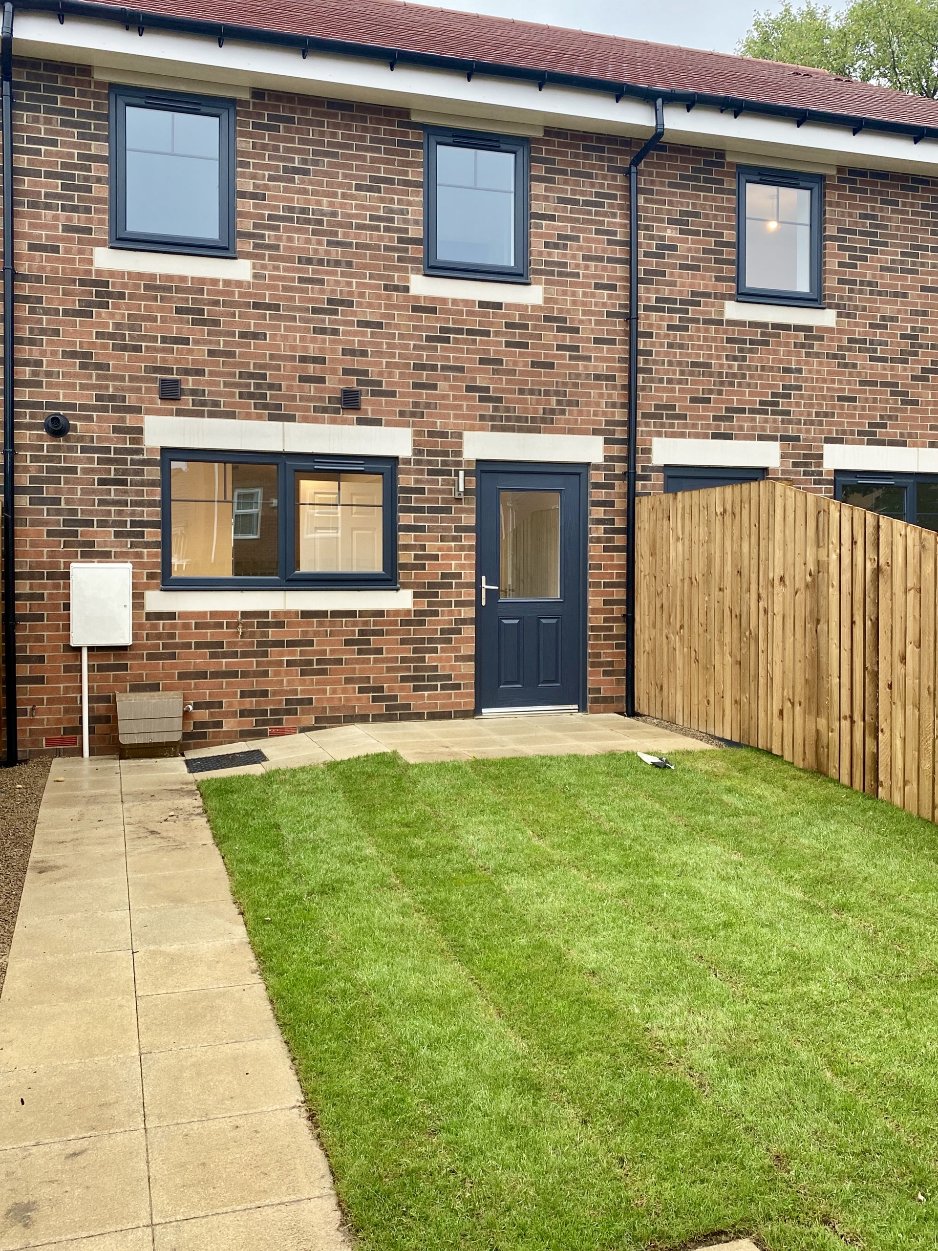 The back of a house with dark brick, navy door and a back garden and pathway to the door.