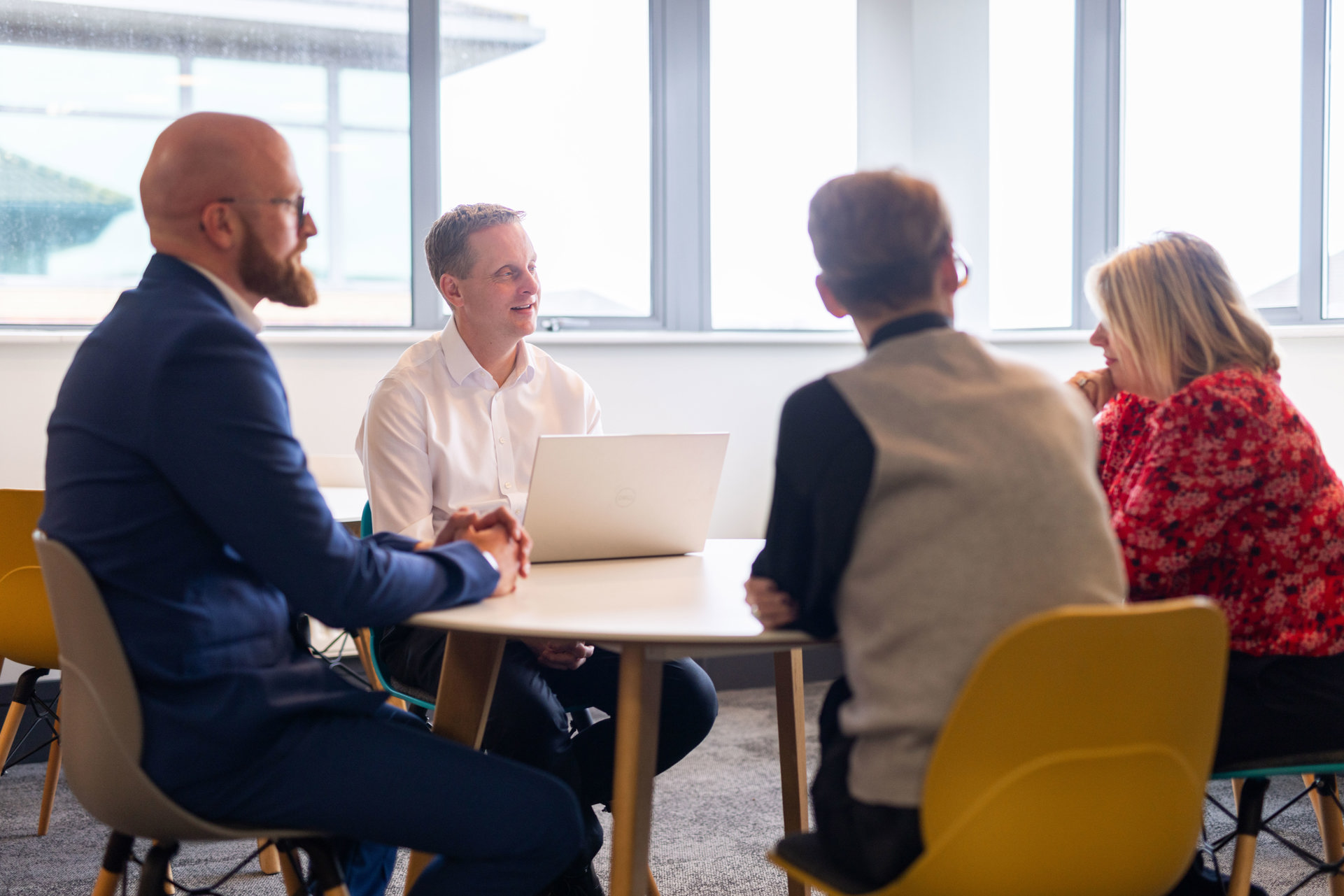 Four people are sitting around a circular table in a modern office space, engaged in a meeting. One man in a white shirt is using a laptop, while the others—two men and one woman—are attentively listening. Large windows in the background let in natural light, creating a bright and professional atmosphere.