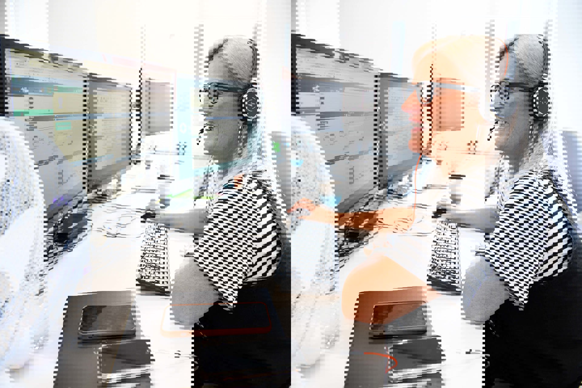  A smiling woman wearing a headset sits at a desk in a bright office, working on a computer with two large monitors. She is wearing glasses and a striped shirt, and appears to be in a customer service or support role. A water bottle, two smartphones, and a coffee mug are visible on the desk. The setting looks professional and friendly.