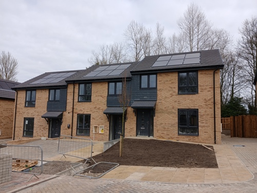 Newly built row of modern two-storey brick houses with solar panels on the roofs. The homes have black doors and windows, small front gardens, and a paved path and driveway in front. A young tree is planted in a central soil bed, and temporary fencing and building materials suggest the final stages of construction.
