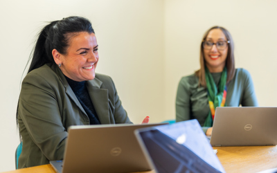 Two people sat next to other other at a table with laptops. The person closest to the camera is in focus and is smiling joyfully.