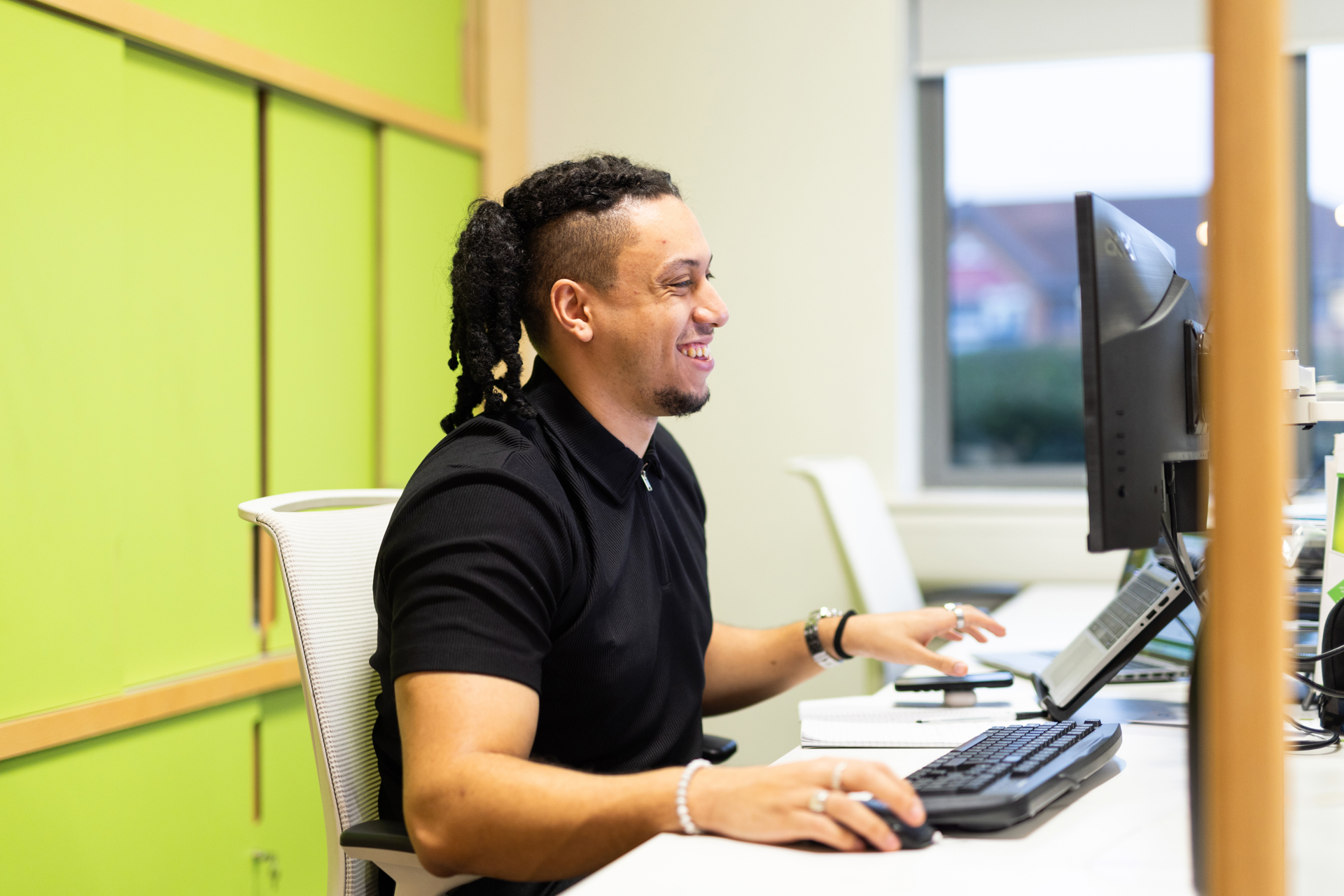 A smiling person sits at a desk in a bright, modern office, working on a desktop computer. They are wearing a black shirt and have long curly hair tied back. The background features a light green wall and large windows letting in natural light. The setting looks welcoming and professional.