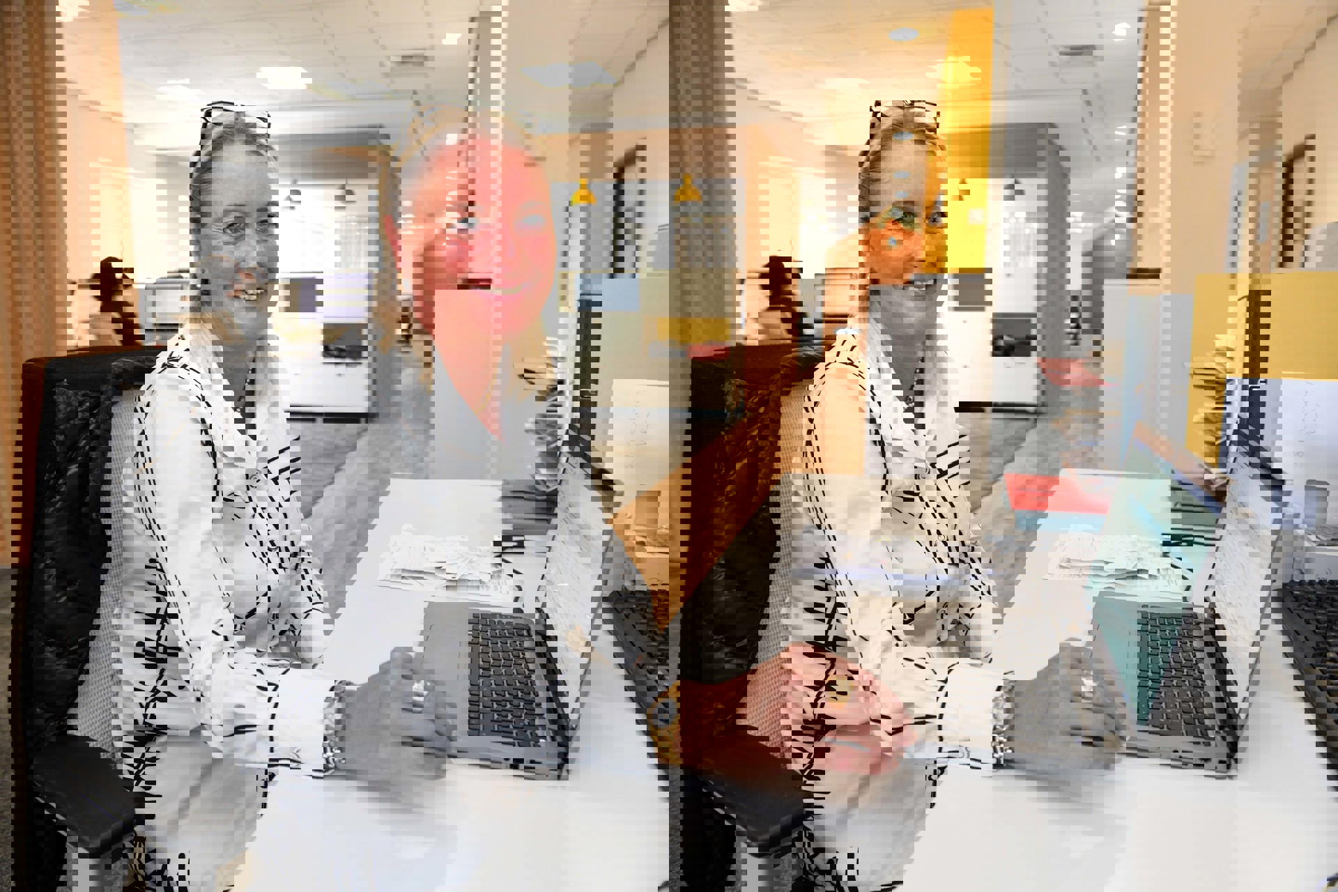  A smiling woman sits at a desk in a bright, modern office space, working on a laptop. She is wearing a white blouse with black patterns and has a pair of glasses resting on her head. Papers and folders are neatly stacked beside her, and the office features a clean, open layout with warm wooden accents