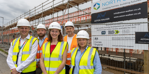 A group of people wearing hard hats and high vis vests stand outside an active building site smiling
