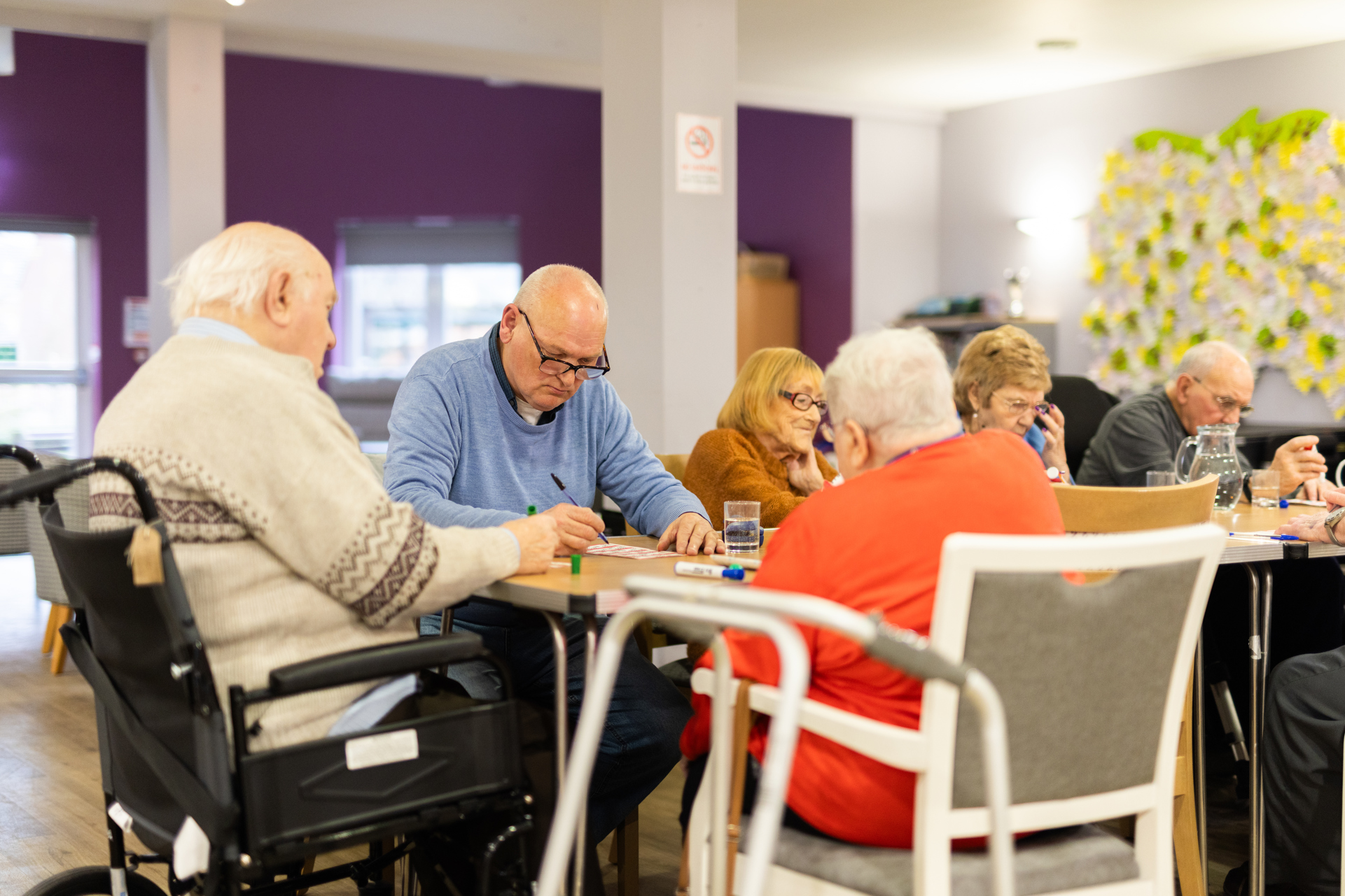 A group of elderly people sat a table in an open, brightly lit room playing bingo.