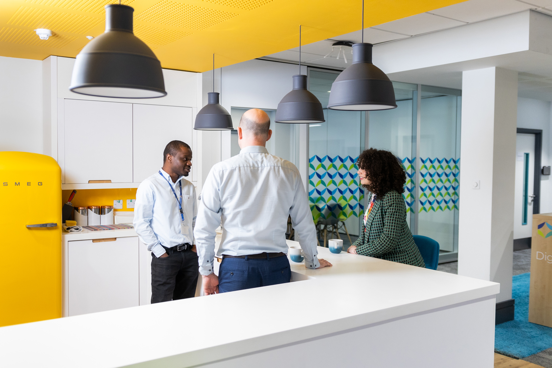 Three people talking in the kitchen area of a modern office. Two are stood and one is sat on a high stool.