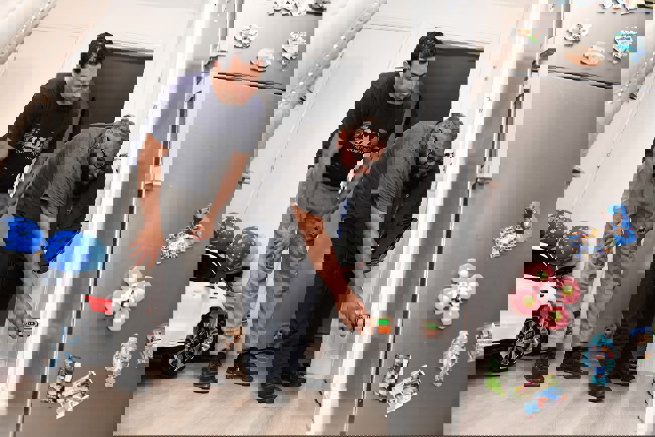 A housing officer uses a handheld device to inspect a wall inside a home, while the resident watches closely. They are in a kitchen area with a fridge covered in magnets and children’s toys visible in the background.  Let me know if you want a version tailored for accessibility compliance or a shorter version for web use.