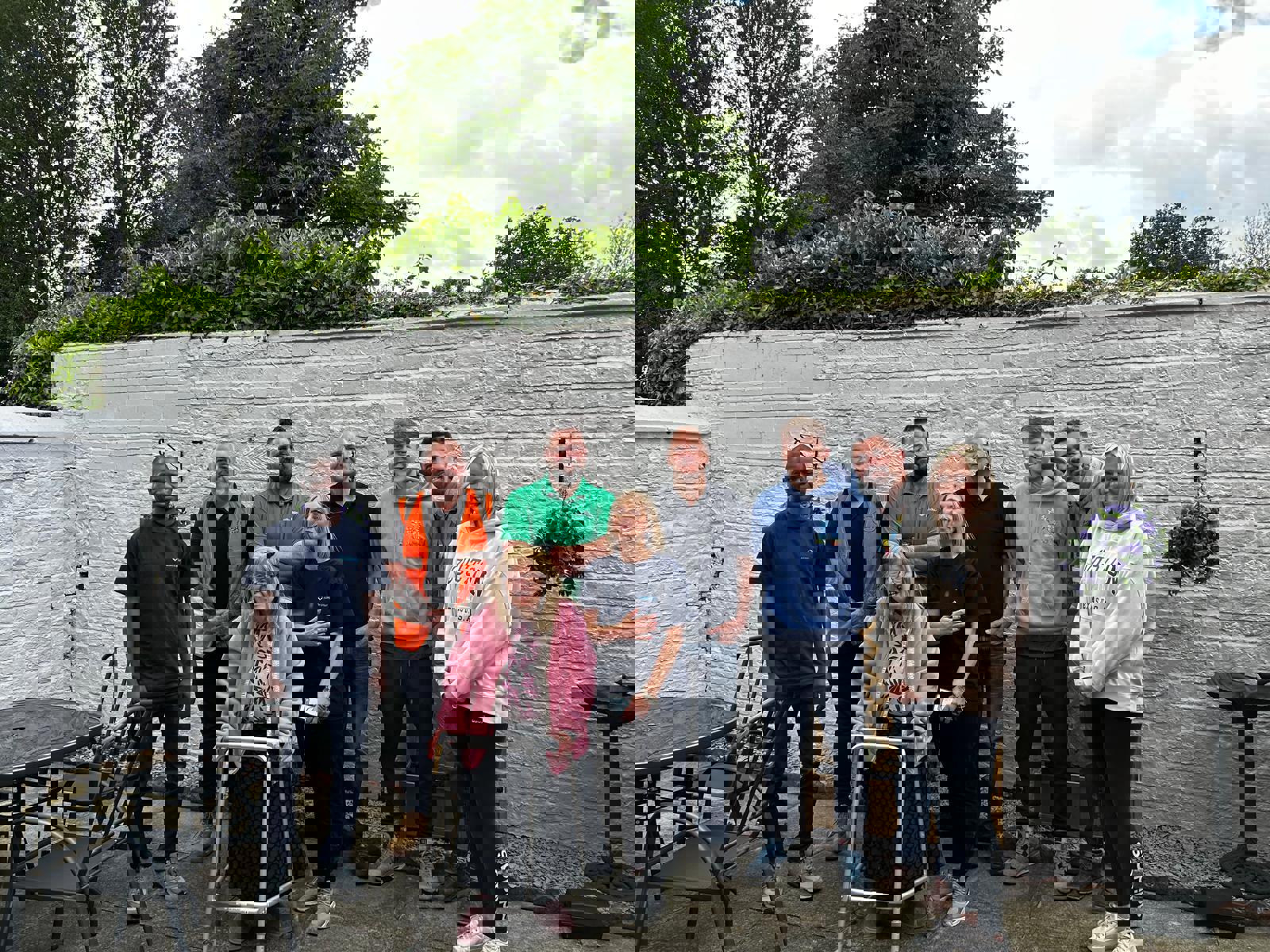 A group of ten people posing outdoors in front of a white brick wall and greenery. The group includes a mix of men and women, some in casual clothing and others in work uniforms or high-visibility vests. A woman using a walking frame is at the front center, smiling alongside the group. The setting appears to be a garden or patio area with outdoor furniture and hanging flowers visible