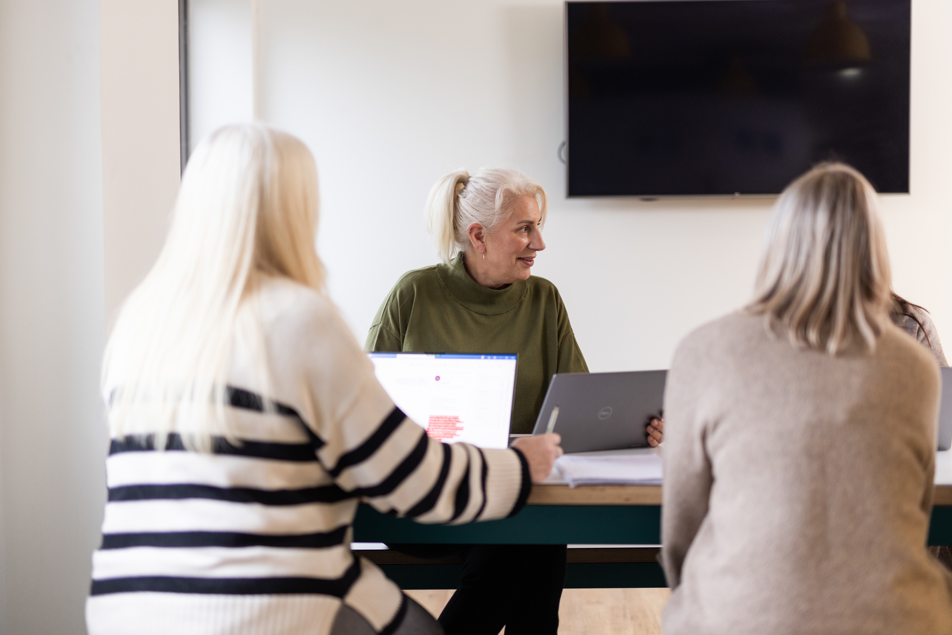 Three people sit at a table in a light room having a work meeting. Two are facing away from the camera. A person between them is facing towards the camera. Their head is turned to the side as if talking to someone out of view. 