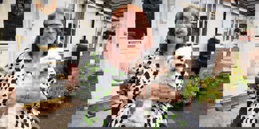 A smiling woman sitting in a wheelchair outside her home, holding a fluffy white dog on her lap and a large mug that reads 'Magnificent Mum.' She is wearing a white dress with green and black spots. Behind her is a white front door with a floral wreath and a garden with colorful potted flowers.