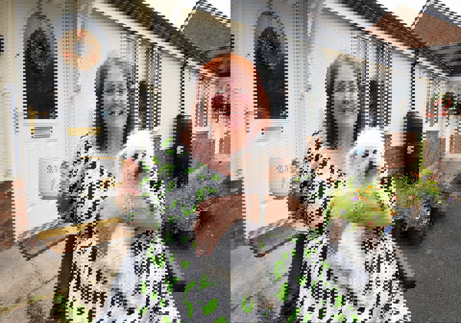 A smiling woman sitting in a wheelchair outside her home, holding a fluffy white dog on her lap and a large mug that reads 'Magnificent Mum.' She is wearing a white dress with green and black spots. Behind her is a white front door with a floral wreath and a garden with colorful potted flowers.